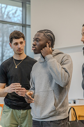 Un homme avec un pull gris qui parle avec à ses côtés : un autre homme tee-shirt noir et chaine dorée